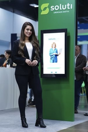 Recepcionista da Turbo ao lado do totem digital no estande da Soluti
